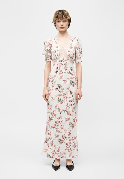 Femme portant une robe longue blanche à fleurs avec des manches bouffantes et un bord en dentelle, debout avec des chaussures noires à bouts pointus sur un fond uni.