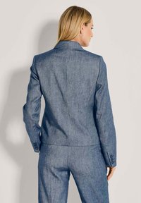 Vrouw met steil blond haar, gekleed in een blauwe denim pak, gezien vanaf de achterkant tegen een lichte achtergrond.