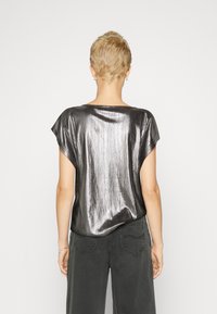 Metallisk silverfärgad topp med korta ärmar, avslappnad passform och en slät, glänsande yta. Har en båtringning och sid sömmar, parat med mörkgrå byxor.