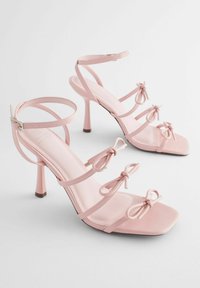 Ein Paar blasser rosa High-Heelsandaletten mit Knöchelriemen, quadratischen Zehen und drei dekorativen Schleifen an den vorderen Riemen jedes Schuhs.