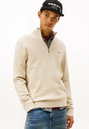 Maglione beige a mezza zip con polsini e orlo a costine, indossato sopra una camicia a quadri, abbinato a jeans blu. Tessuto morbido e strutturato.