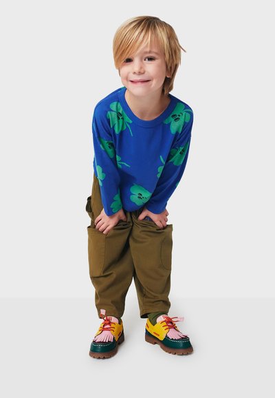 Blaues Langarmhemd mit grünen Blattformen, olivgrüne Hose und mehrfarbige Schuhe mit pinken Akzenten und einem strukturierten Design.