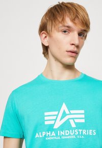Türkises Baumwoll-T-Shirt mit Rundhalsausschnitt, das über einen weißen Logo-Text und ein grafisches Design verfügt. Der Stoff wirkt weich und leicht.