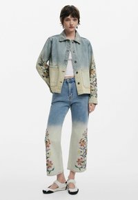 Jeansjacke mit mehrfarbigem Blumenstickerei an den Ärmeln, kombiniert mit hellblauen Jeans mit Blumenmustern und schwarz-weißen Schuhen.