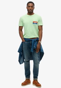Camiseta verde claro de cuello redondo con un logo de bolsillo azul, rojo y blanco, combinada con jeans azules, una camisa de mezclilla atada a la cintura y botas marrones.