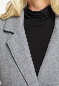 Blazer en feutre gris avec des revers aiguisés sur un col roulé noir. Tissu texturé avec une finition lisse. Pas de quincaillerie ou de motifs visibles.