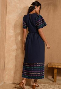 Abito midi blu navy con bordo e maniche scallop colorate, elastico in vita, dettaglio con apertura a chiave sul retro, indossato con sandali con tacco a blocco marroni.