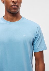 Világoskék pamut póló kerek nyakkal és rövid ujjakkal, amelyen a mellrészen egy finom fehér logó található. Sima tapintású.