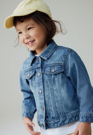 Lächelndes junges Kind mit einer hellgelben Mütze und einer blauen Jeansjacke, das vor einem schlichten hellgrauen Hintergrund steht.