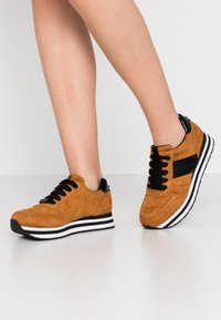 Zapatillas de ante marrón con acentos negros, con cordones negros y una suela blanca a rayas. Punta redondeada y material texturizado para mayor agarre.