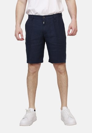 Uomo con pantaloncini blu navy fino al ginocchio, t-shirt bianca e scarpe da ginnastica bianche, in piedi con le braccia rilassate lungo i fianchi.