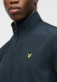 Κοντινό πλάνο ενός ατόμου που φοράει σκούρο μπλε μπουφάν με φερμουάρ και μικρό κίτρινο πουλί στο αριστερό στήθος.