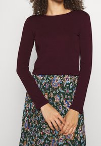 Kvinna iklädd en åtsittande långärmad mörklila topp och en grön blommönstrad kjol, med händerna låsta framför kroppen vid midjehöjd.