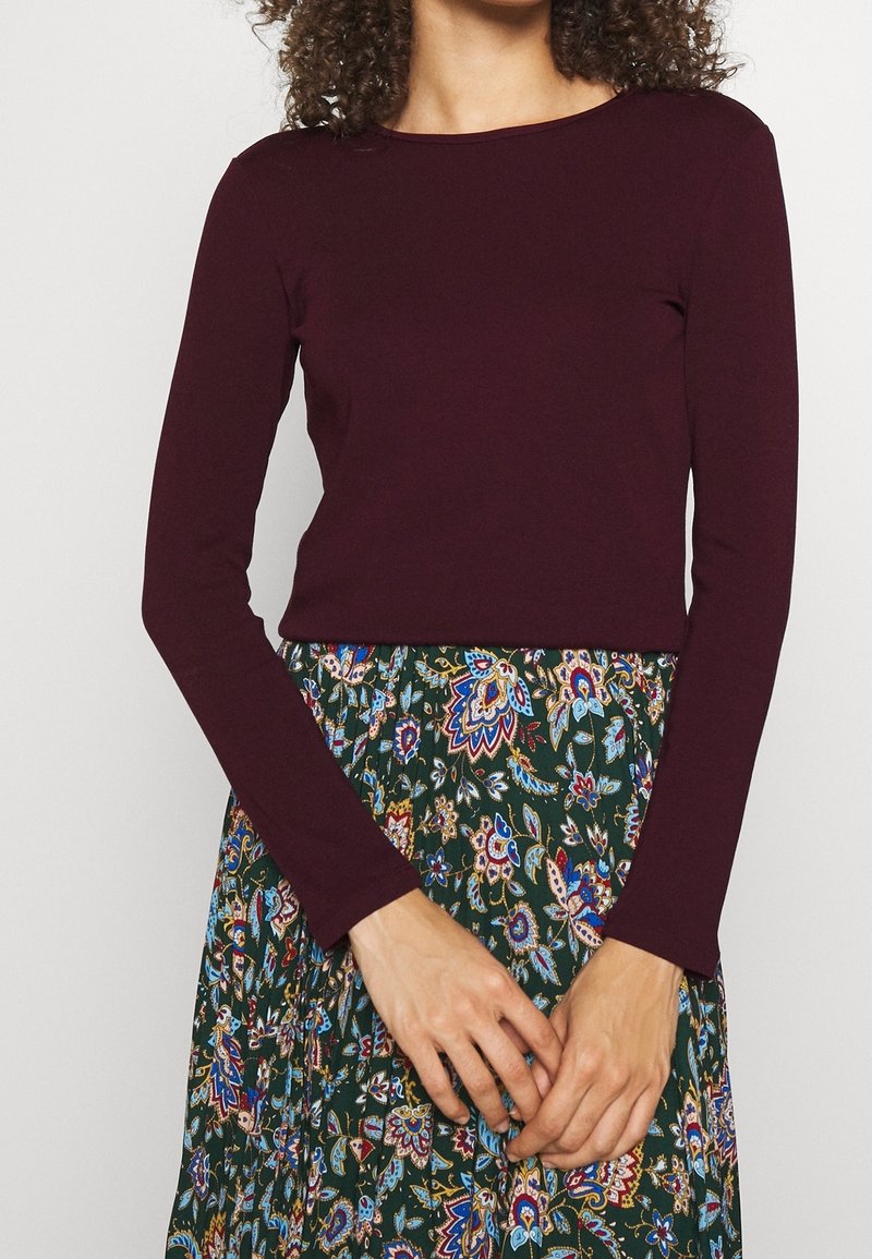 Kobieta ubrana w dopasowaną, długą, ciemnofioletową bluzkę z długim rękawem i zieloną spódnicę w kwiatowy wzór, z dłońmi złożonymi przed sobą na wysokości talii.