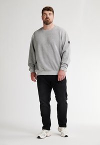 Grijze crewneck sweatshirt met gestructureerde stof, geribde manchetten en onderrand, gecombineerd met zwarte spijkerbroeken en witte sneakers. Model staat tegen een neutrale achtergrond.