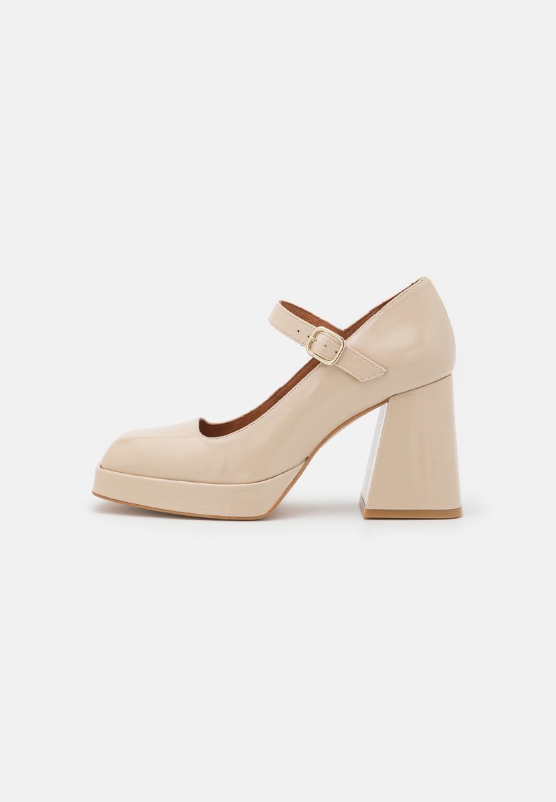 Beige lackskinn Mary Jane-skon med kraftig klack och ankelrem med ett guldfärgat spänne, visad från sidan på vit bakgrund.