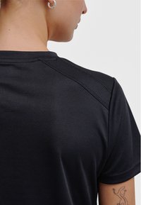 Czarna sportowa koszulka z gładkim, teksturowanym materiałem, krótkimi rękawami i widocznymi szwami na ramieniu. Zawiera wzór tatuażu na ramieniu.