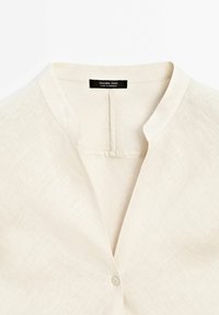Beige linned bluse med kinesisk krave, knaplukning og en tekstureret stof. Mærket er synligt indeni kraven.