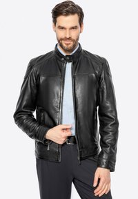 Giacca di pelle nera con colletto rialzato, zip frontale e tasche laterali; presenta una texture liscia e un design aderente. Indossata sopra una camicia azzurra chiara.