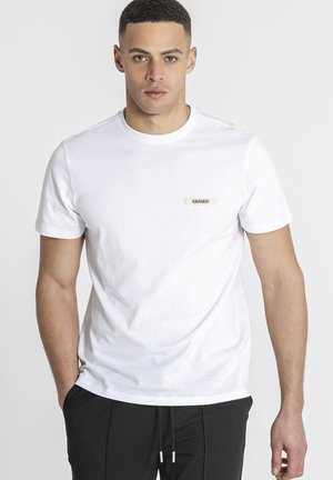 Ung mand iført en helt hvid T-shirt med teksten "Kavanagh" på brystet og sorte snor-bukser, stående mod en hvid baggrund.