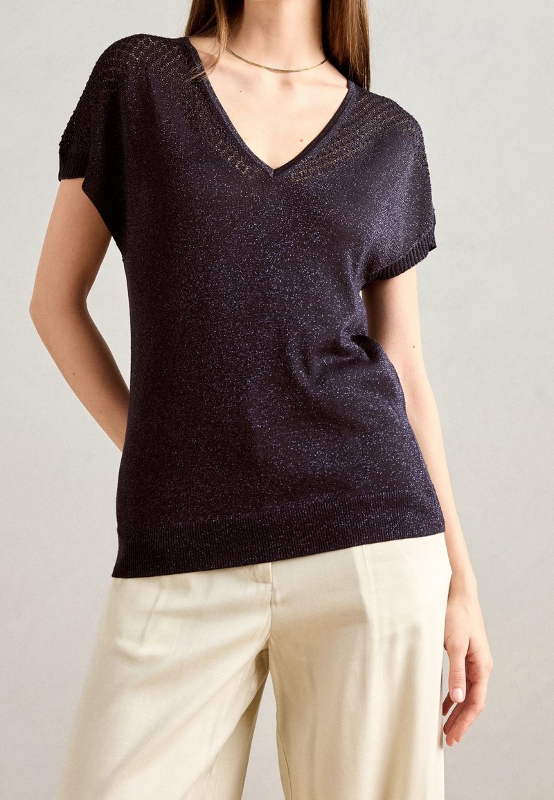 Femme portant un haut à manches courtes, col en V scintillant de couleur sombre, associé à un pantalon beige clair, sur un fond uni.