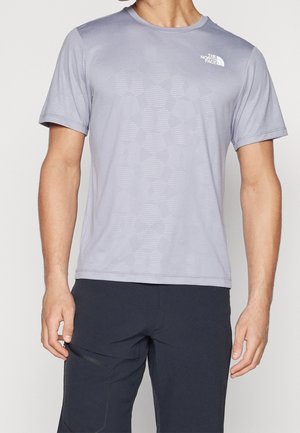 Homme portant un t-shirt North Face à manches courtes violet clair avec un motif géométrique subtil et un pantalon noir avec une poche zippée sur la jambe gauche.