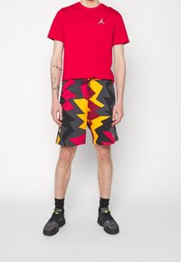Hombre vistiendo una camiseta roja y pantalones cortos con un patrón en zigzag vibrante en negro, rojo, amarillo y gris, de pie frente a un fondo blanco.