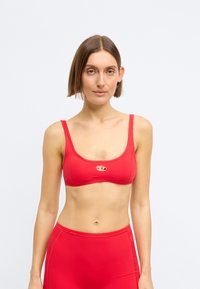 Νεαρή γυναίκα με κοντά καστανά μαλλιά, που φοράει κόκκινο αθλητικό σουτιέν και κόκκινο σλιπ με ψηλή μέση, πάνω σε απλό ανοιχτό φόντο.