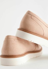 Halvány rózsaszín velúr slip-on cipők fehér gumi talppal és barna díszvarrással. Alacsony profilú kialakítás sima textúrával és minimális részletezéssel.