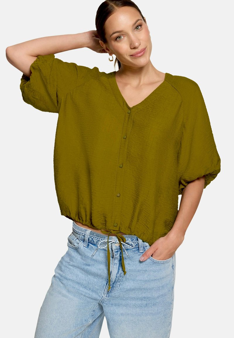 Femme portant un chemisier olive boutonné avec un cordon de serrage à l'ourlet et un jean bleu clair, posant avec un bras derrière la tête.