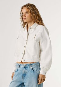 Jauna sieviete ar cirtainiem blondiem matiem valkā baltu īsu ar pogām aizsprādzētu jaku un gaiši zilas augsta vidukļa džinsas, skatās pa kreisi.