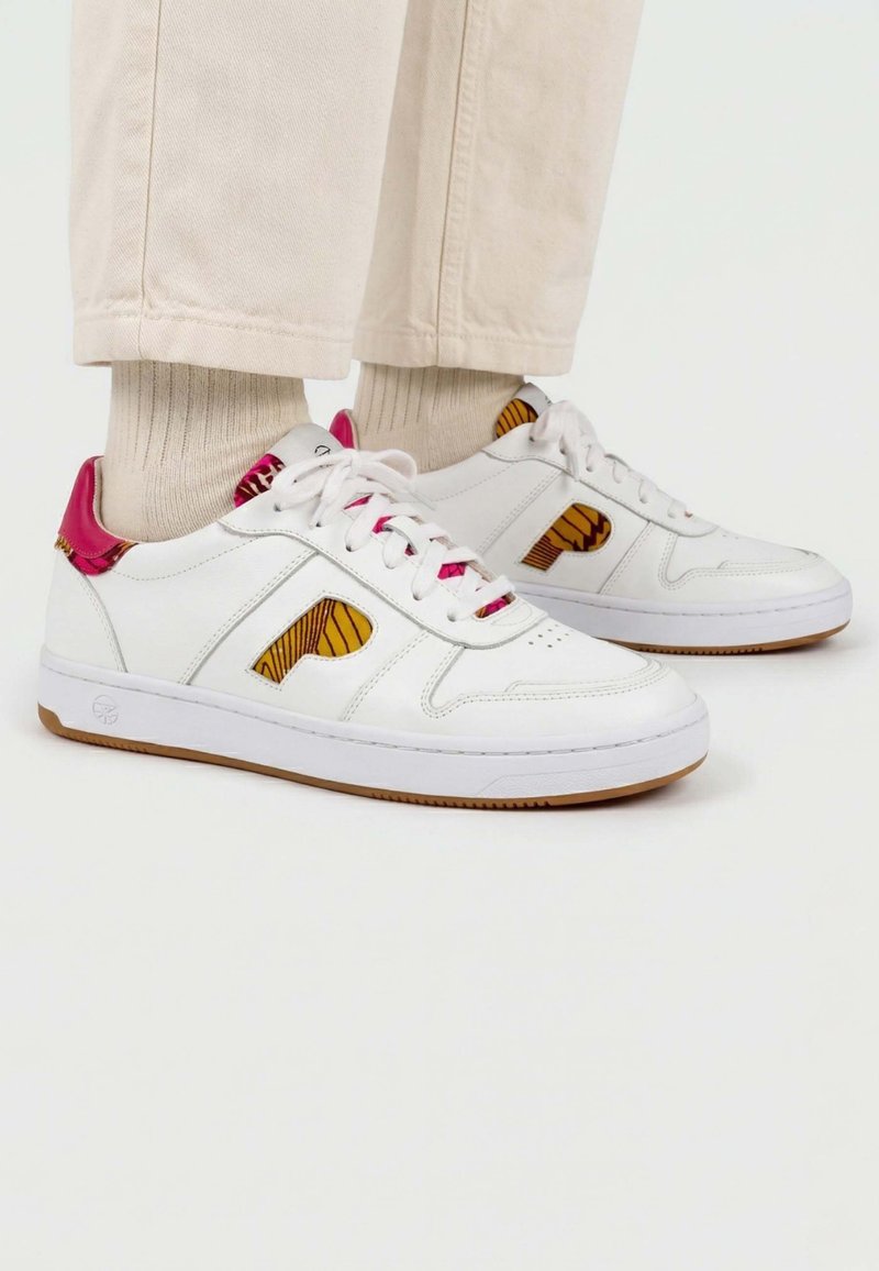 Baskets blanches en cuir avec des détails latéraux marron à motifs, des accents roses au talon et à la languette, portées avec des chaussettes beige et un pantalon crème.