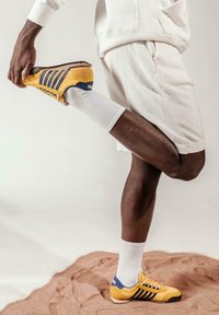 Une personne portant des shorts blancs et des chaussettes étire une jambe vers l'arrière, tenant une chaussure de sport jaune et bleue contre un fond uni.