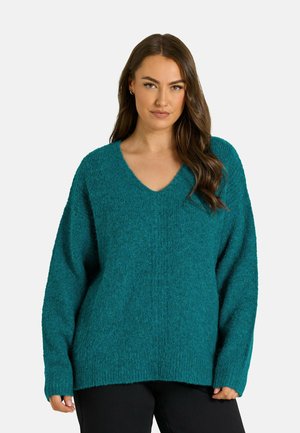 Femme aux longs cheveux bruns portant un pull à col en V duveteux couleur sarcelle et un pantalon noir sur un fond clair uni.