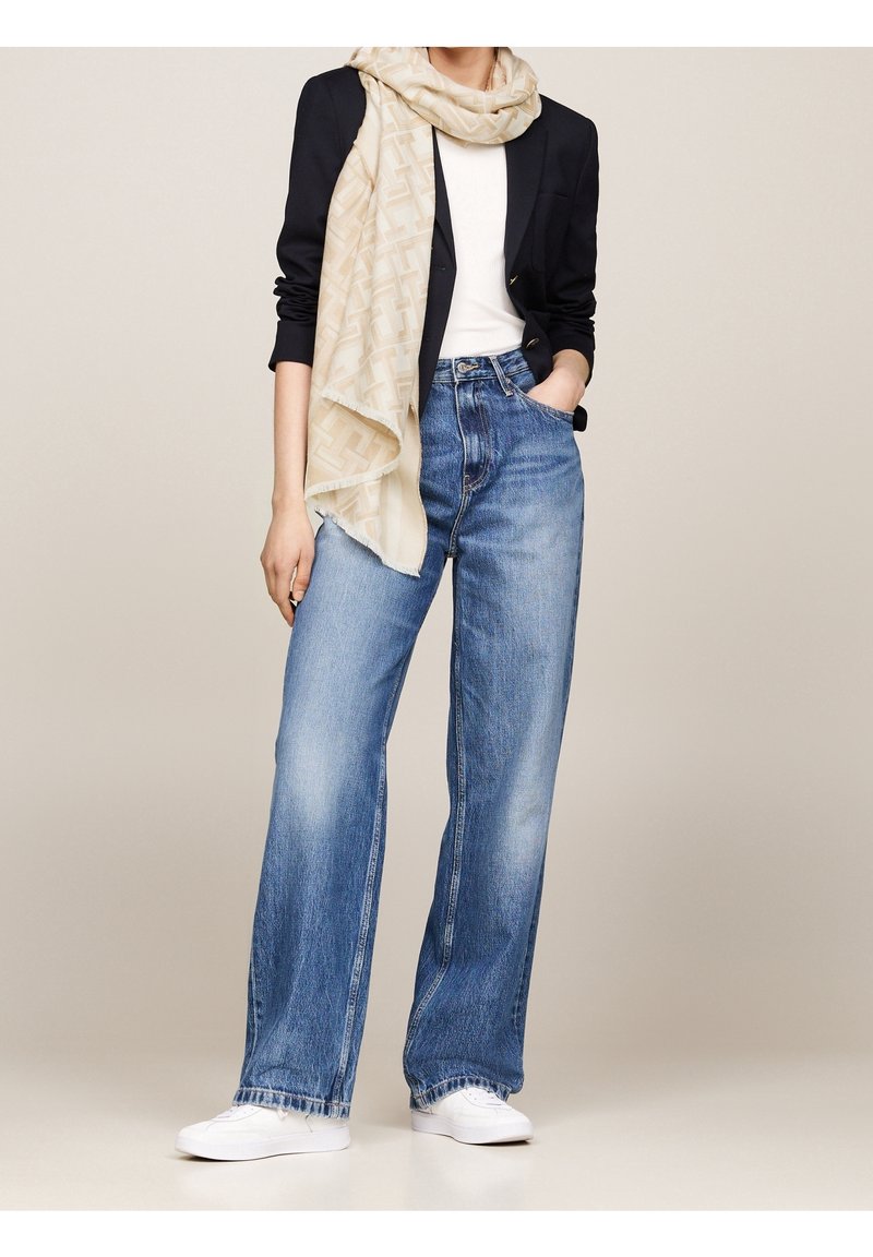 Femme portant un jean bleu à jambes larges, des baskets blanches, un haut blanc, un blazer noir et une écharpe beige à motifs, main dans la poche, debout devant un fond neutre.