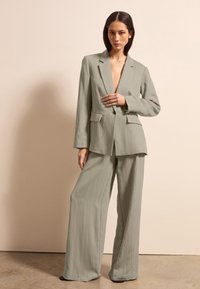 Lichtgroen tweedelig pak gemaakt van gestructureerde stof. De blazer heeft een gelapd schouder en twee voorzakken. De broek is wijd gesneden.