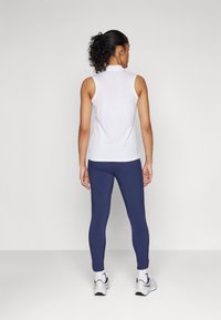 Valkoinen hihaton toppi yhdistettynä istuviin tummansinihin legginsseihin. Asusteessa on sileät materiaalit ja minimalistinen muotoilu. Urheilukengät näkyvissä.