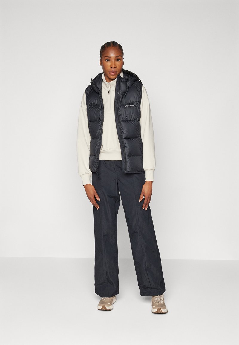 Czarna kamizelka puchowa z kapturem noszona na kremowej bluzie, w połączeniu z luźnymi ciemnymi spodniami i beżowymi sneakersami. Gładka tekstura, funkcjonalny design.