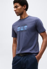 Man draagt een marineblauw T-shirt met opdruk en donkere broek, staat met zijn handen in de zakken en kijkt zijwaarts tegen een effen achtergrond.