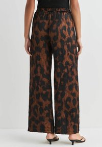 Vrouw in losse, gestructureerde wijde broek met een bruin en zwart luipaardpatroon, gecombineerd met zwarte hoge sandalen en een zwart topje.