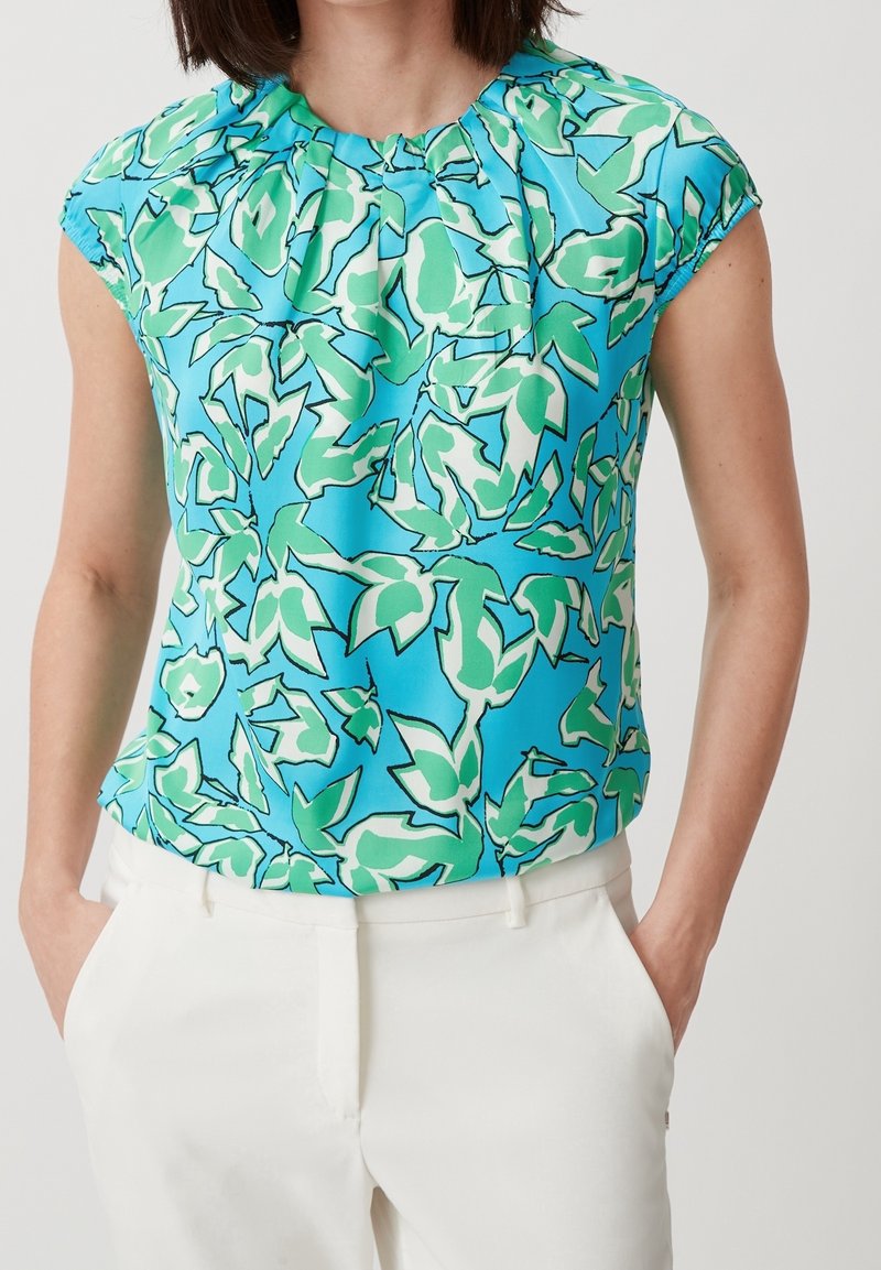 Femme portant un haut bleu sans manches avec un motif abstrait de feuilles vertes et un pantalon blanc, les mains dans les poches.