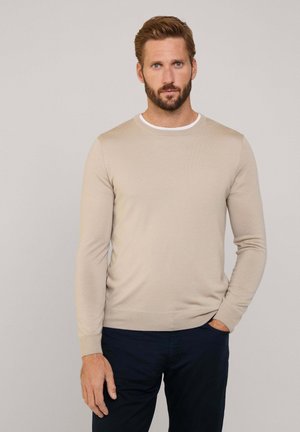 Suéter beige de manga larga con cuello redondo y ribetes blancos. Hecho de una tela suave, con forma ajustada y puños acanalados. Combinado con pantalones oscuros.