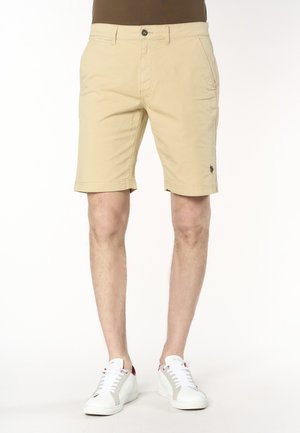 Pantaloncini beige alla lunghezza del ginocchio con tasche laterali indossati da una persona, abbinati a scarpe da ginnastica bianche e una maglietta marrone.