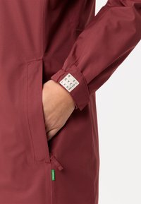 Giacca impermeabile color marrone con finitura texturizzata, caratterizzata da una tasca laterale, polsini regolabili e una piccola etichetta verde vicino all'orlo.