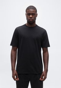 Νέος άνδρας που φοράει απλό μαύρο κοντομάνικο μπλουζάκι και μαύρο παντελόνι, στέκεται μπροστά σε ανοιχτό γκρι φόντο, κοιτάζοντας προς τα εμπρός.