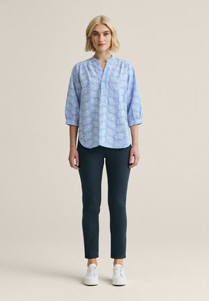 Blond kvinde iført en lyseblå blomstret bluse med 3/4 ærmer, sorte slanke bukser og hvide sneakers, stående mod en ensfarvet baggrund.