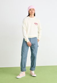 Cream knit sweater with red "Dickies" logo, light blue cargo pants, and pink high-top sneakers. Model wears a pink beanie, standing on green floor.