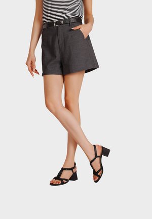 Donna con sandali neri con tacco a blocco e cinturino, shorts grigio scuro con cintura e top a righe bianche e nere, in piedi con le gambe incrociate.
