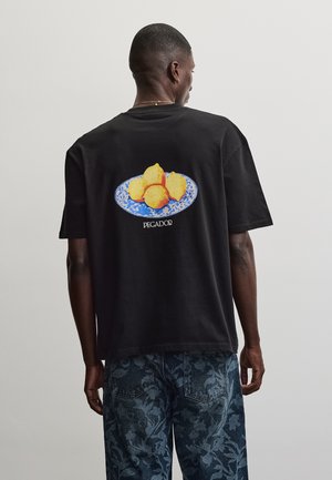 Uomo che indossa una maglietta nera con una targhetta blu che tiene quattro limoni e la scritta "PEGADOR" sulla schiena, abbinata a pantaloni blu con motivo floreale.
