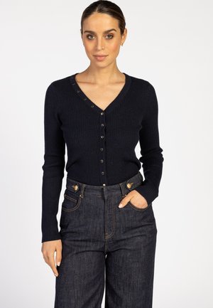 Vrouw in een donkerblauwe ribgebreide cardigan met knopen en een hoge taille donkere spijkerbroek, staand met één hand in haar zak.